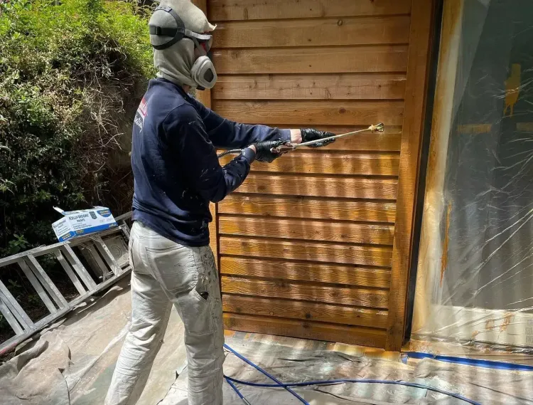 Person in a respirator pressure-washes a wooden wall beside a window outdoors.