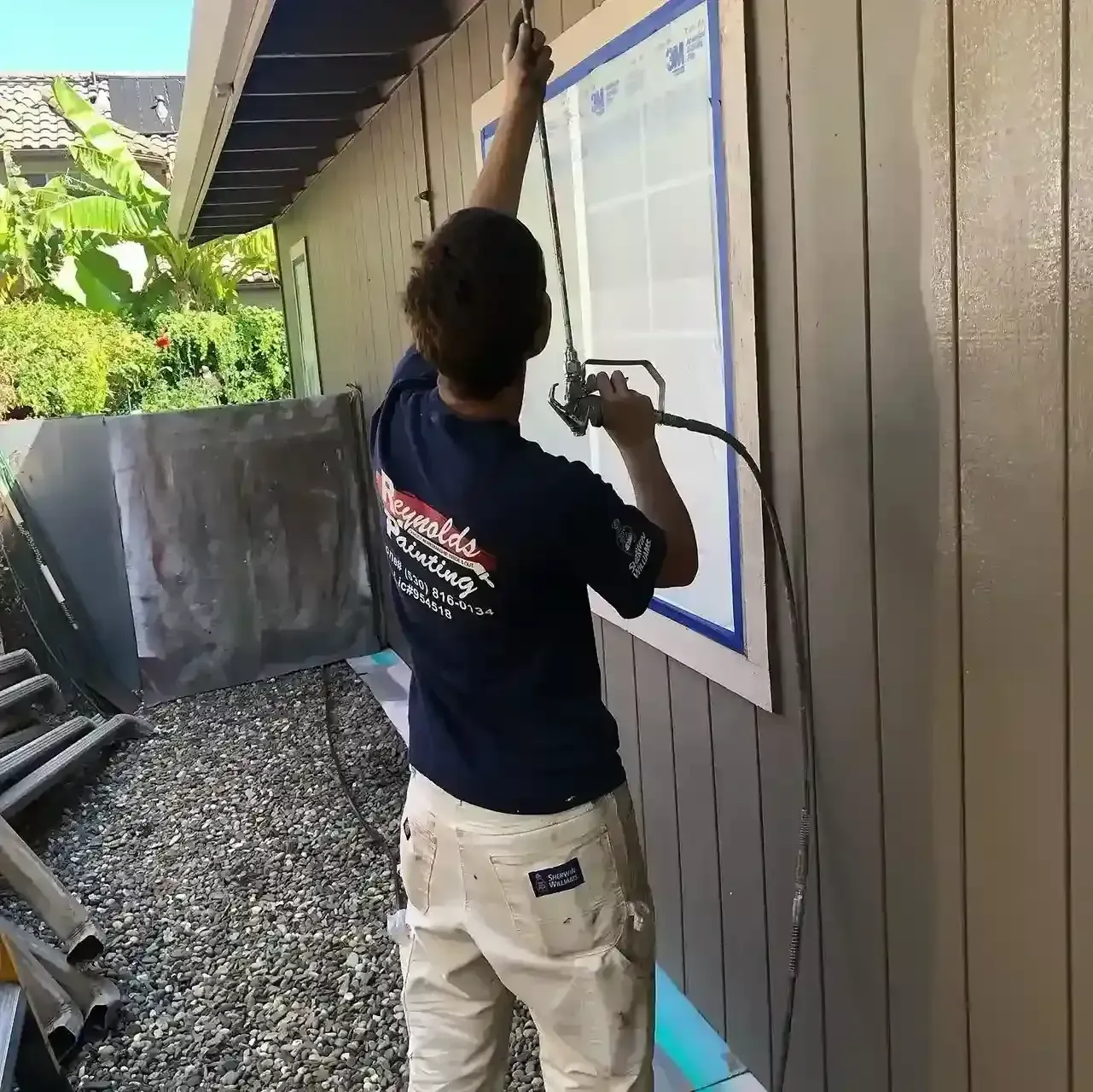 Person spray-painting a house exterior window trim with blue painter’s tape.