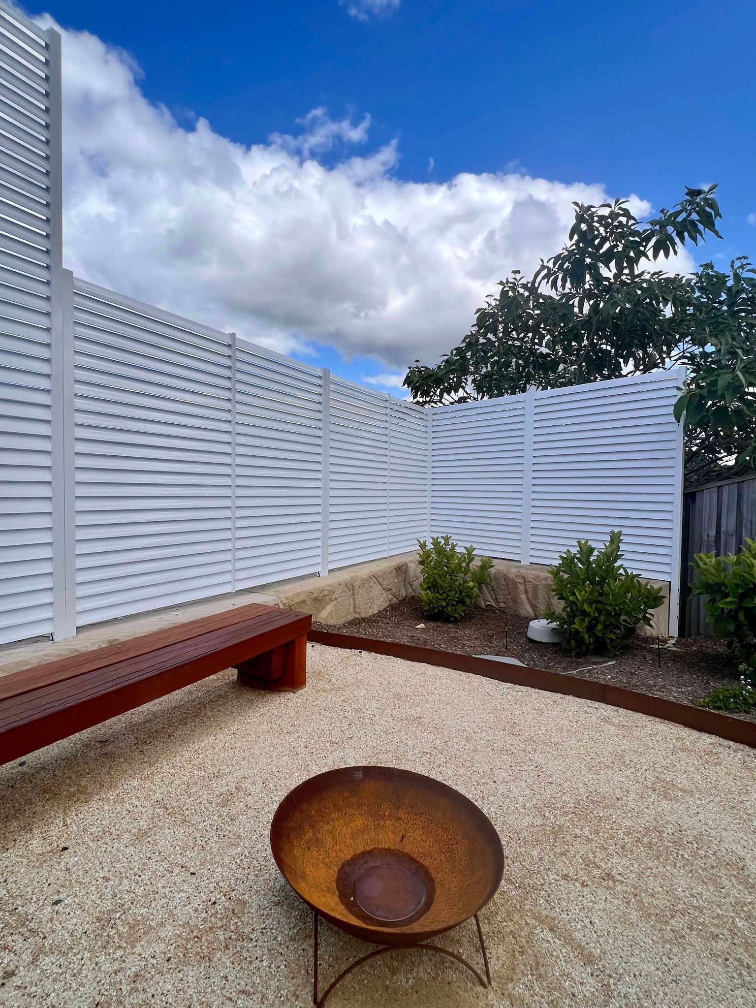 A Rusty Metal Fire Pit Sits on A Gravel Patio Beside a Wooden Bench — Coastline Glass Pool Fencing in Old Bar, NSW