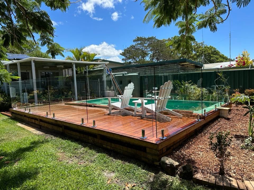 A Swimming Pool with A Wooden Deck and Chairs Around It — Coastline Glass Pool Fencing in Cundletown, NSW