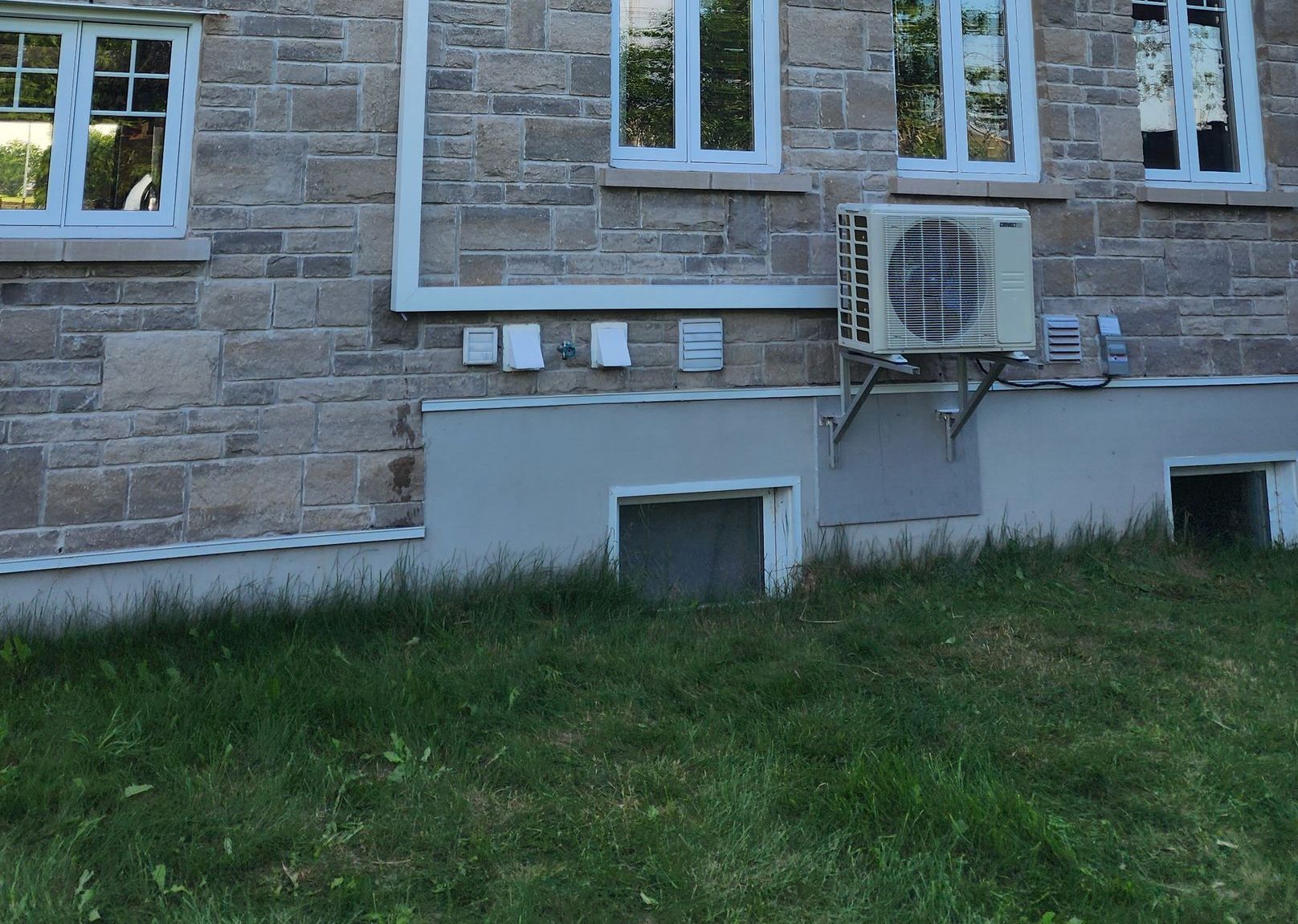 Le mur extérieur d'une maison en pierre avec un système de chauffage, de ventilation et de climatisation, plusieurs fenêtres et des bouches d'aération au niveau du sol, au-dessus d'une pelouse.