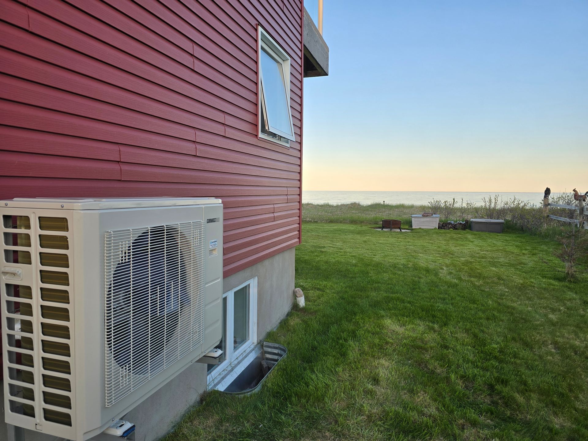 Une pompe à chaleur est installée à l'extérieur d'une maison aux murs rouges, dotée d'une fenêtre au sous-sol, donnant sur une cour herbeuse en bord de mer au crépuscule.