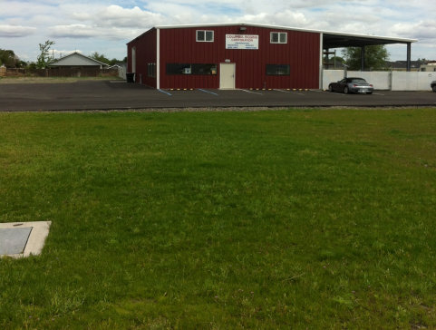 Red building with a sign, set behind a grassy field and paved lot under a cloudy sky.