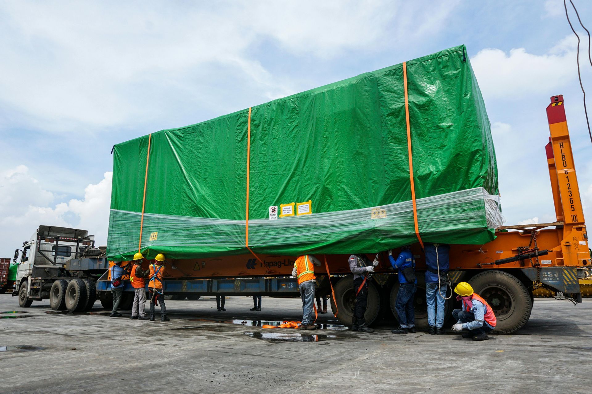 Workers securing a large green tarped load on a truck in an open industrial yard