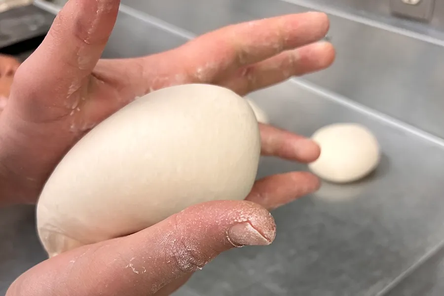 Hand holding a smooth, pale dough ball on a floured surface, with another dough ball in the background