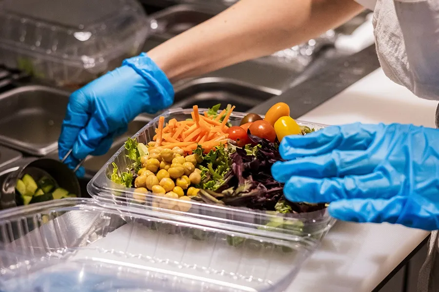 Gloved hands arranging a salad in a clear takeout container at a kitchen counter