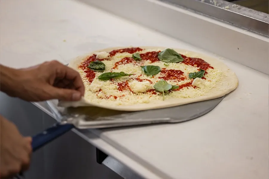 Hands sliding a pizza onto a peel in a kitchen, topped with cheese, tomato sauce, and basil
