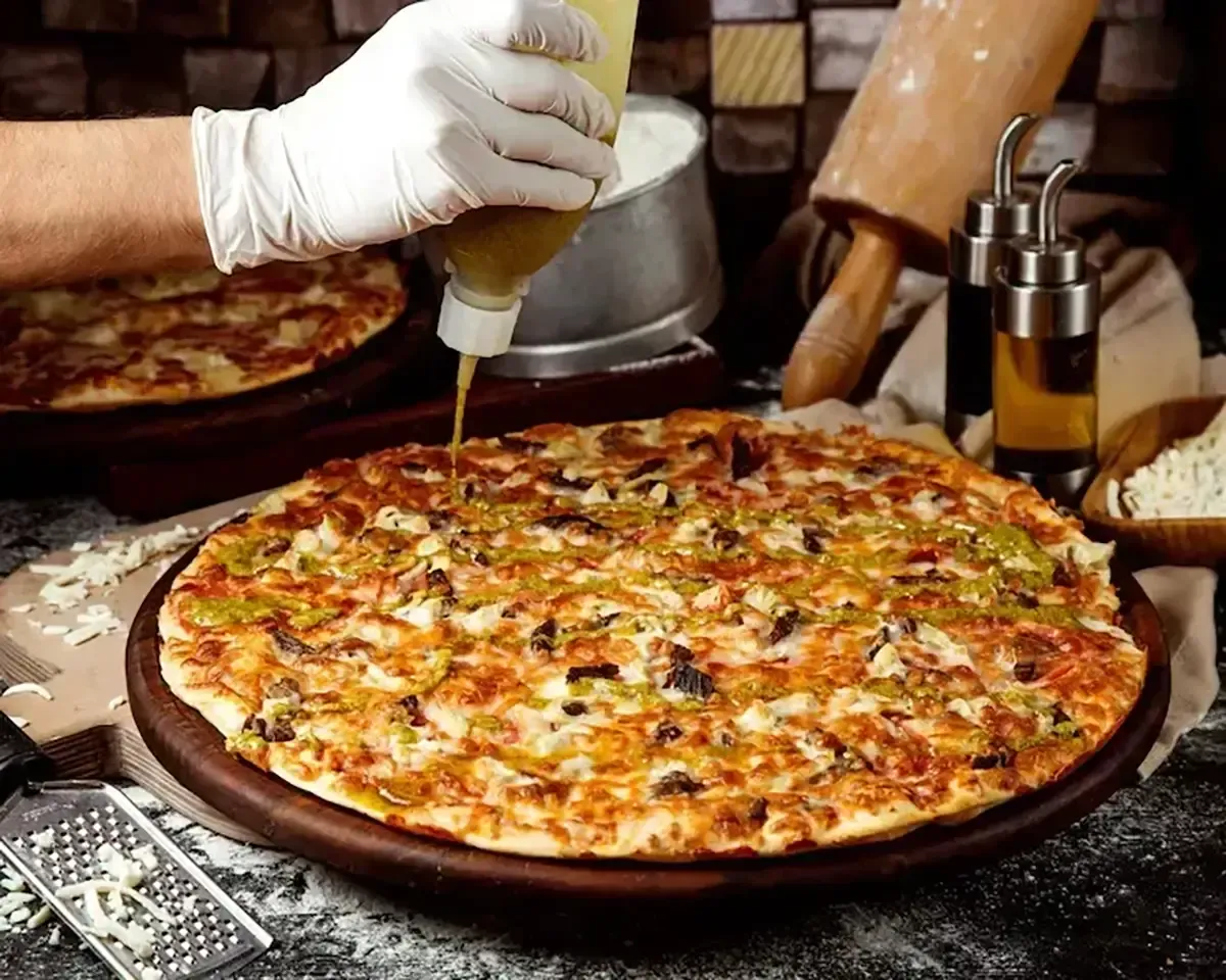 Gloved hand drizzling sauce on a large pizza on a counter, with utensils and ingredients nearby