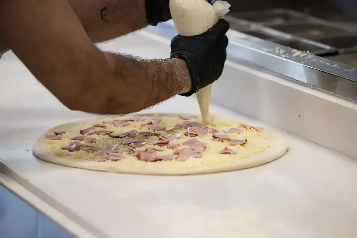 Gloved worker piping sauce onto a pizza on a prep counter
