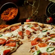 Fresh veggie pizza with tomatoes, olives, and mushrooms on a wooden board, with blurred spices in the background