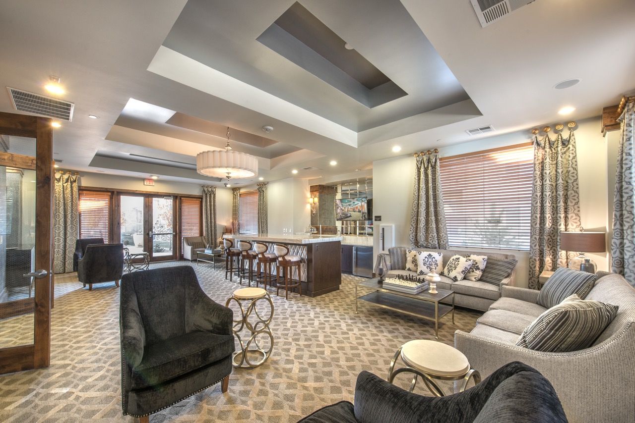 Indoor resident lounge with sofas, armchairs, a bar, and large windows.