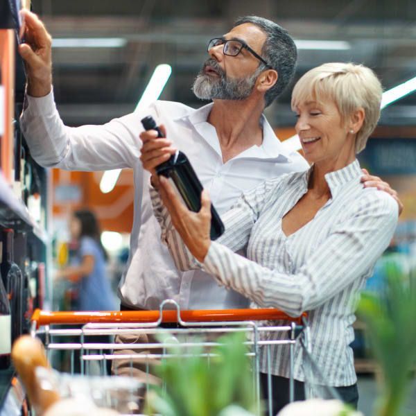 A couple shops in a store, smiling as they examine a dark glass bottle while pushing a cart.