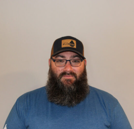 Man with a beard, glasses, and black cap in a blue shirt against a plain wall.