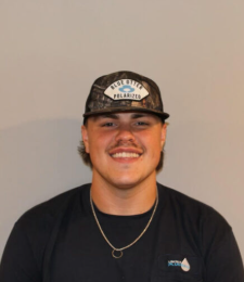 Smiling person in a black shirt and camo cap against a plain beige wall