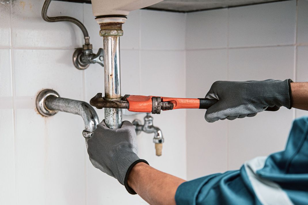 Gloved plumber tightening orange pipe under a sink with a wrench