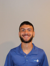 Smiling man in a blue polo shirt standing against a plain beige wall.