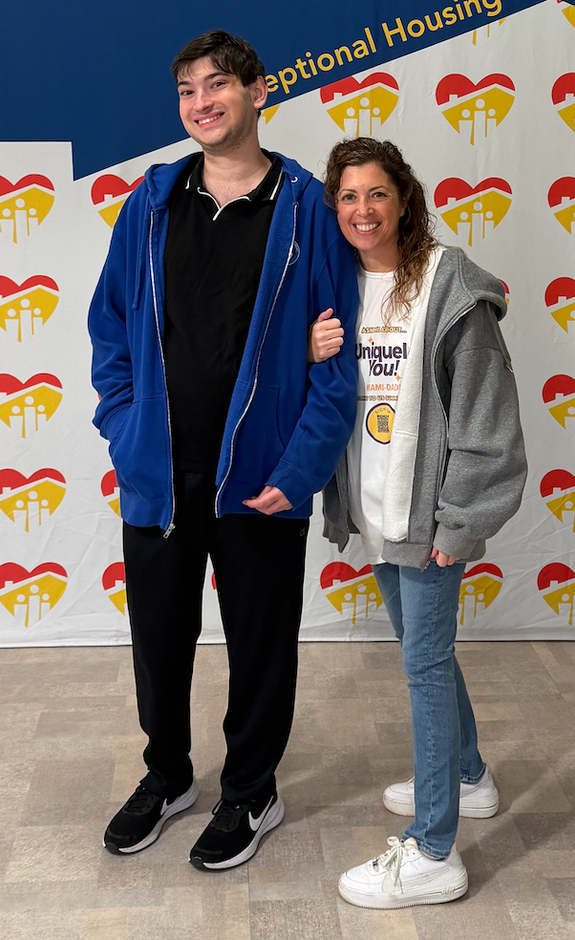 Two people stand side-by-side smiling in front of a backdrop featuring a repeating heart-shaped logo.