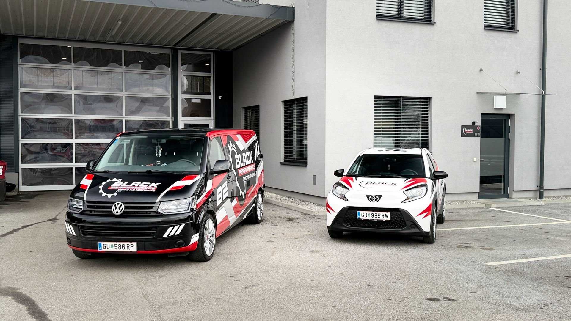 Zwei Rallyeautos, eines schwarz-rot und eines weiß-rot, parkten vor einer Garage auf einem grauen Parkplatz.