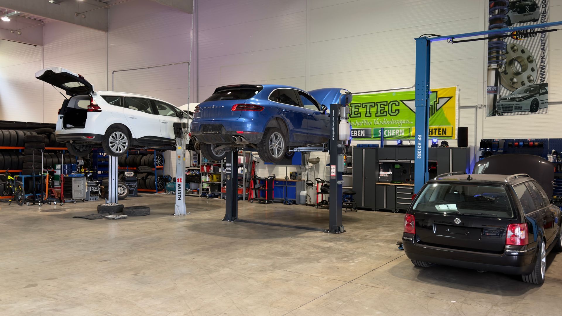 Autowerkstatt mit drei Autos auf Hebebühnen und einem schwarzen Schrägheckwagen davor.