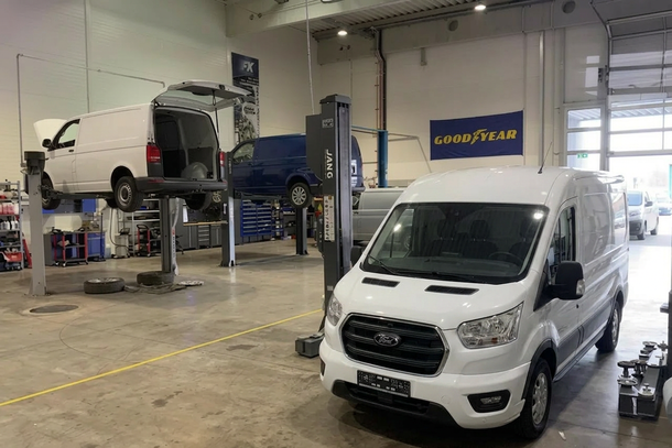 Autowerkstatt mit einem weißen Lieferwagen davor und einem blauen Fahrzeug auf einer Hebebühne im Hintergrund