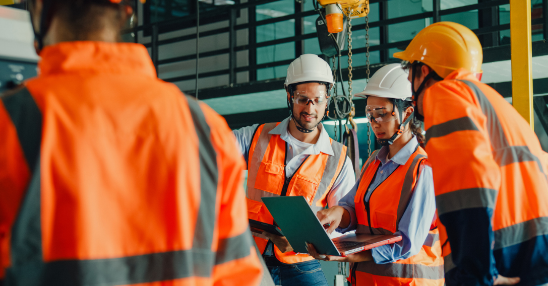 Operários da construção civil, usando coletes de segurança laranja e capacetes, analisam plantas em um canteiro de obras.