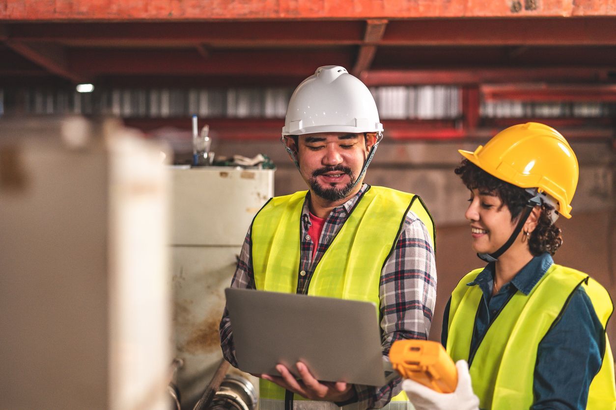 Dois operários com capacetes e coletes de segurança analisando um laptop em um canteiro de obras.