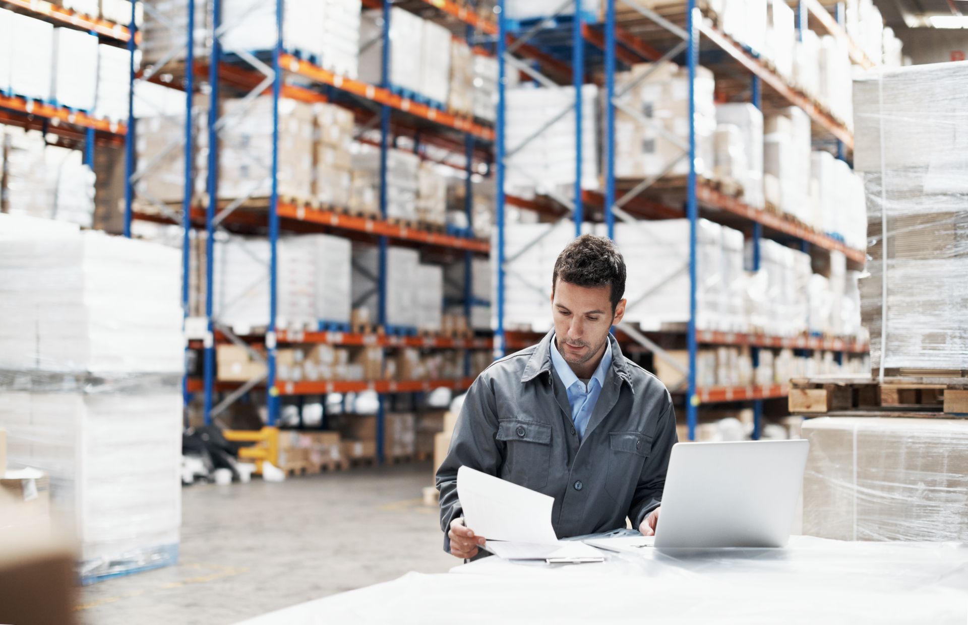 Homem revisando documentos em uma mesa de armazém ao lado de um laptop e prateleiras de produtos embalados.