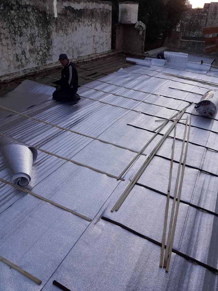 Un trabajador arrodillado en un tejado instala láminas aislantes plateadas y listones de bambú.