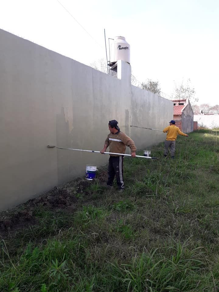 Dos trabajadores pintan una larga pared exterior junto a la hierba crecida