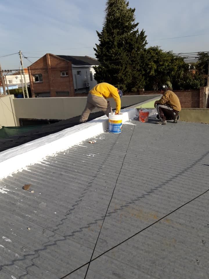 Dos trabajadores reparando un tejado, uno con una camisa amarilla cerca de un cubo de pintura