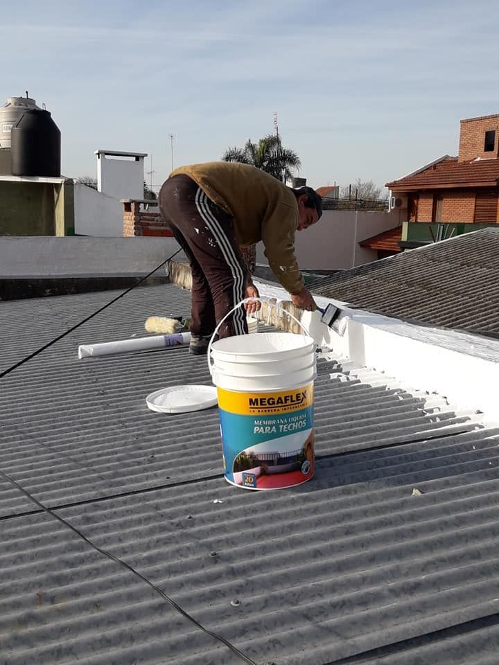 Un trabajador pinta un techo de chapa ondulada junto a un cubo blanco de pintura en una azotea.