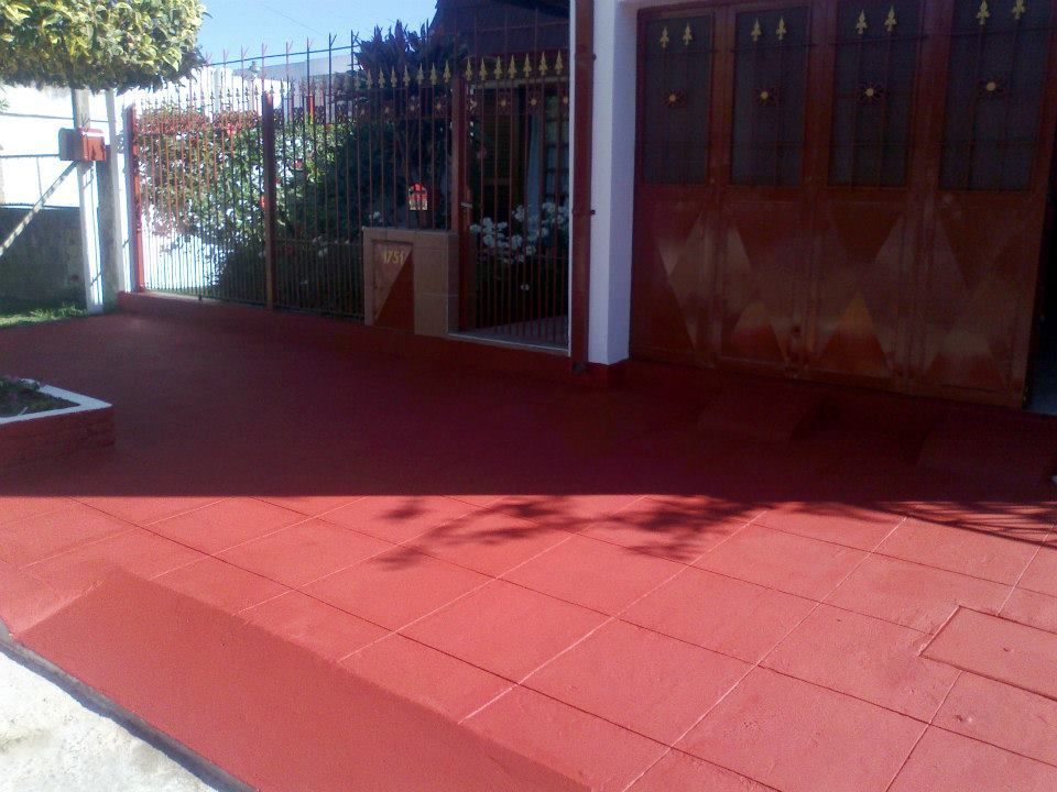 Patio con baldosas rojas bañado por la luz del sol junto a una casa con verja de metal y puertas de madera.