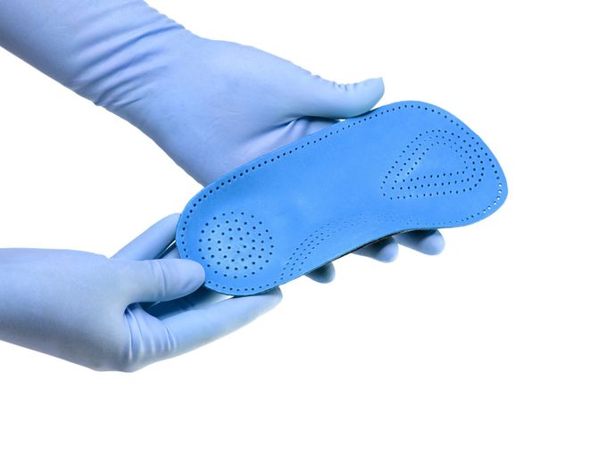 Mani con guanti in nitrile blu che reggono una soletta per scarpe in schiuma blu perforata su sfondo bianco.