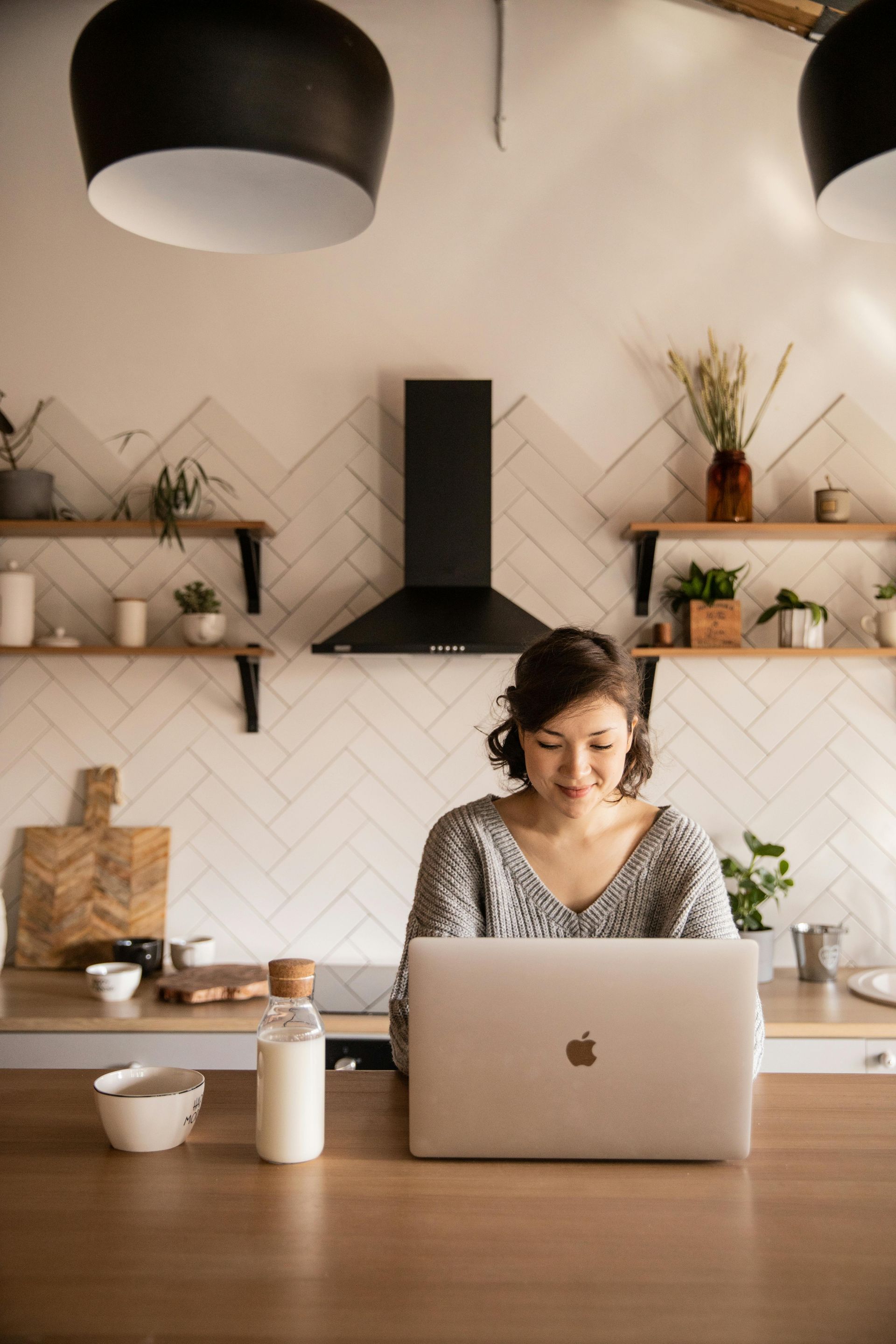 Mulher na cozinha trabalhando em um laptop. Ela sorri, com leite e uma tigela por perto.
