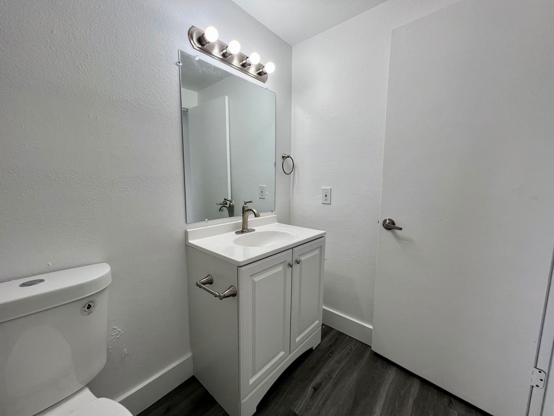 Small white bathroom with vanity, mirror lights, toilet, and an open door.
