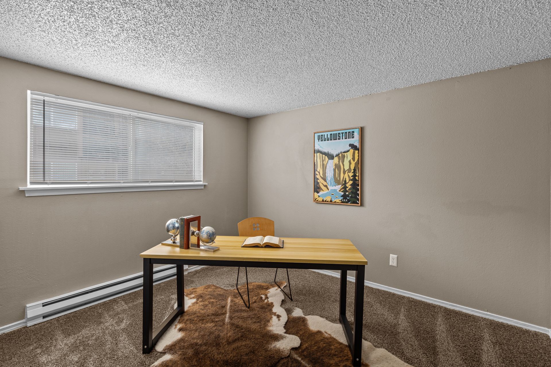 Empty office with a desk, chair, window blinds, beige walls, and a wall painting.