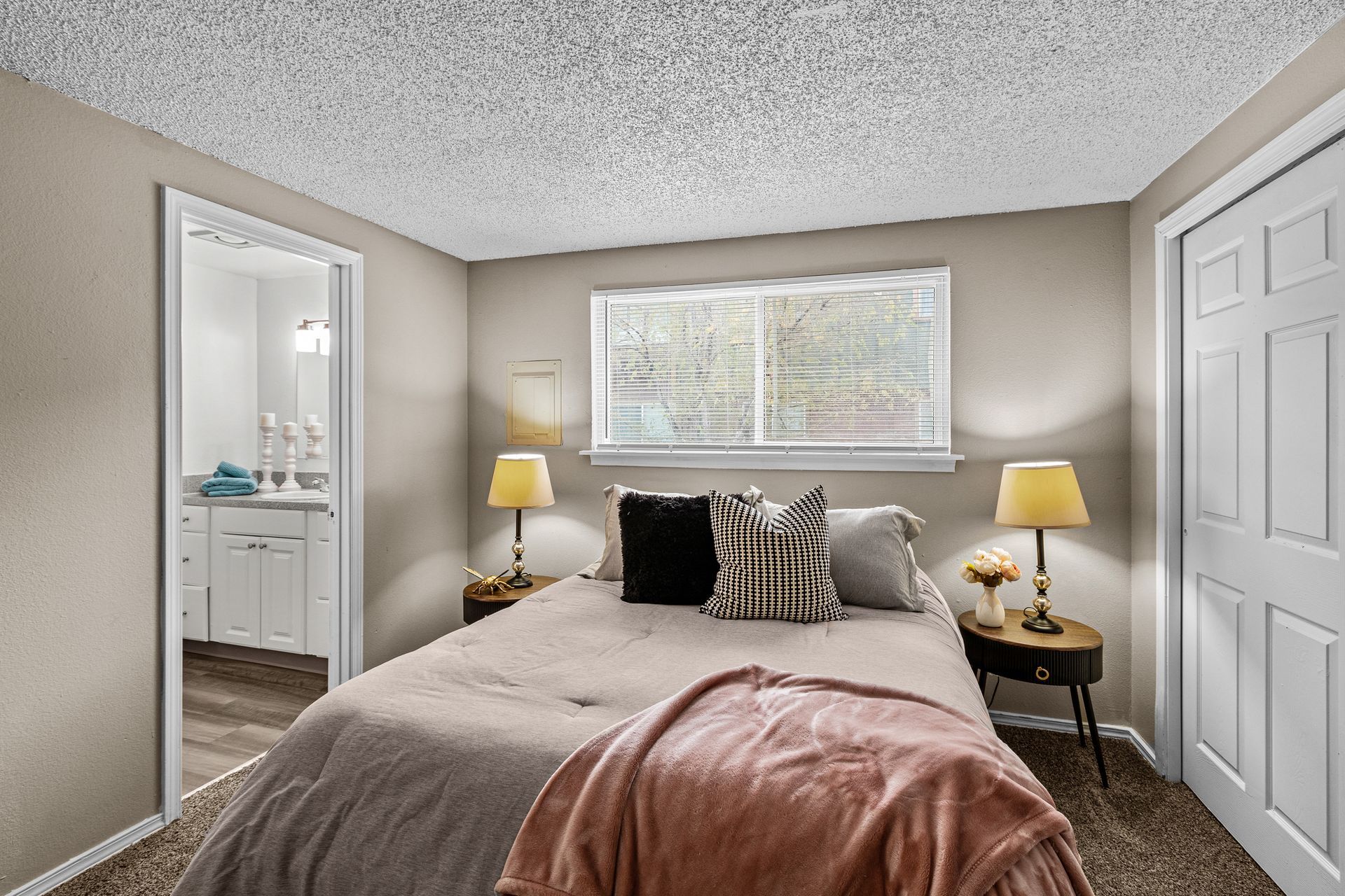 Cozy bedroom with a made bed, bedside lamps, window blinds, and carpeted floor