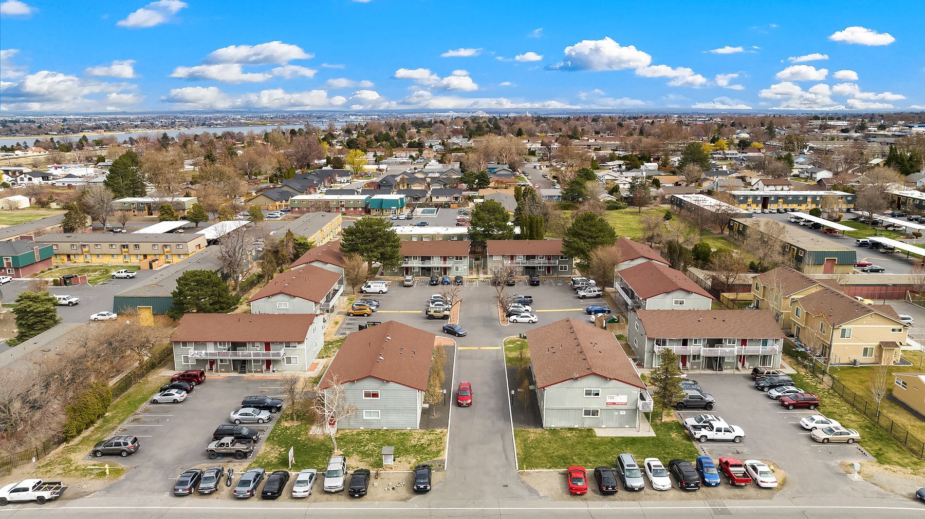 Aerial view of a suburban apartment complex with parked cars, red roofs, and a clear blue sky.