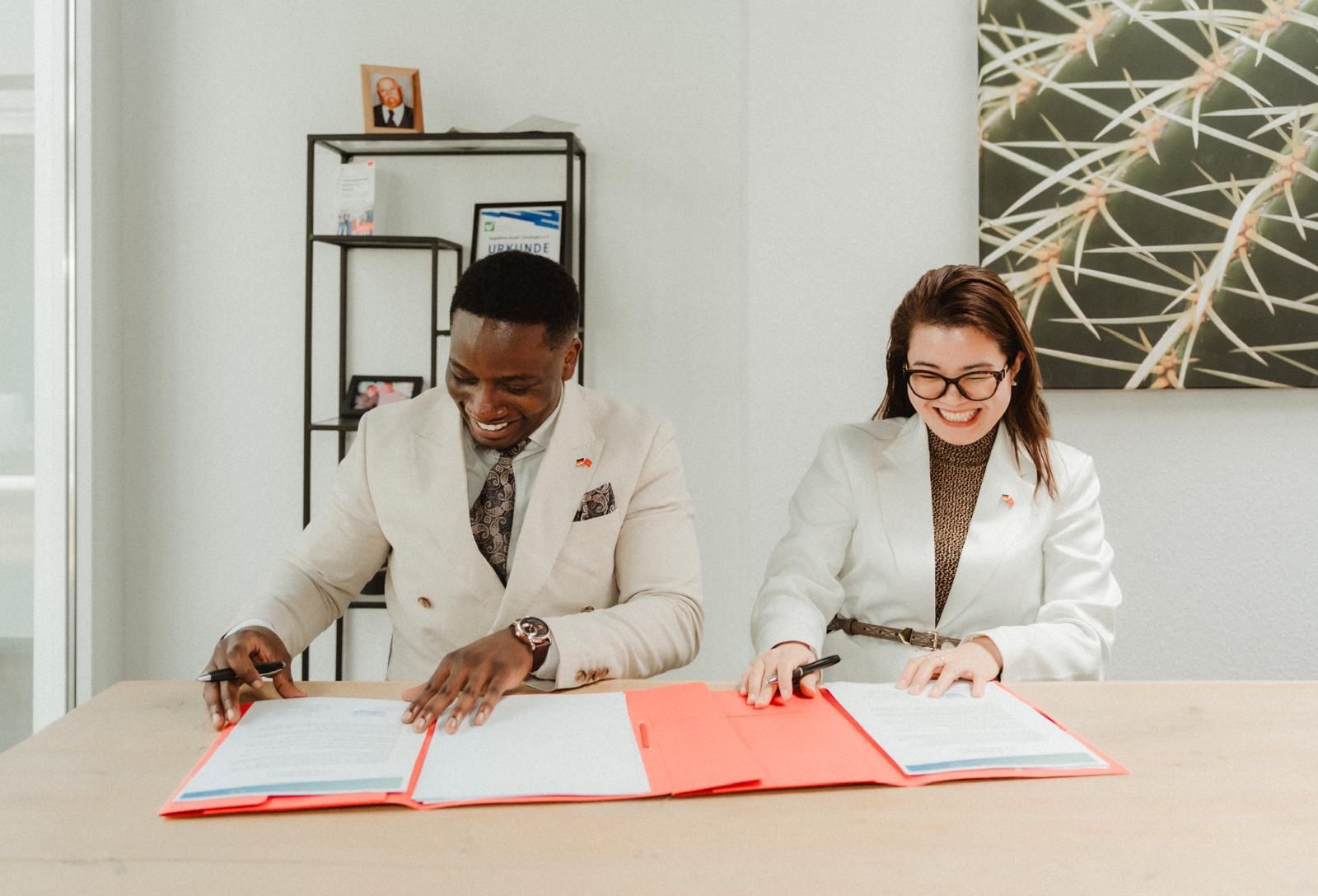 Zwei Personen unterschreiben an einem Tisch Dokumente. Beide lächeln und tragen Blazer.