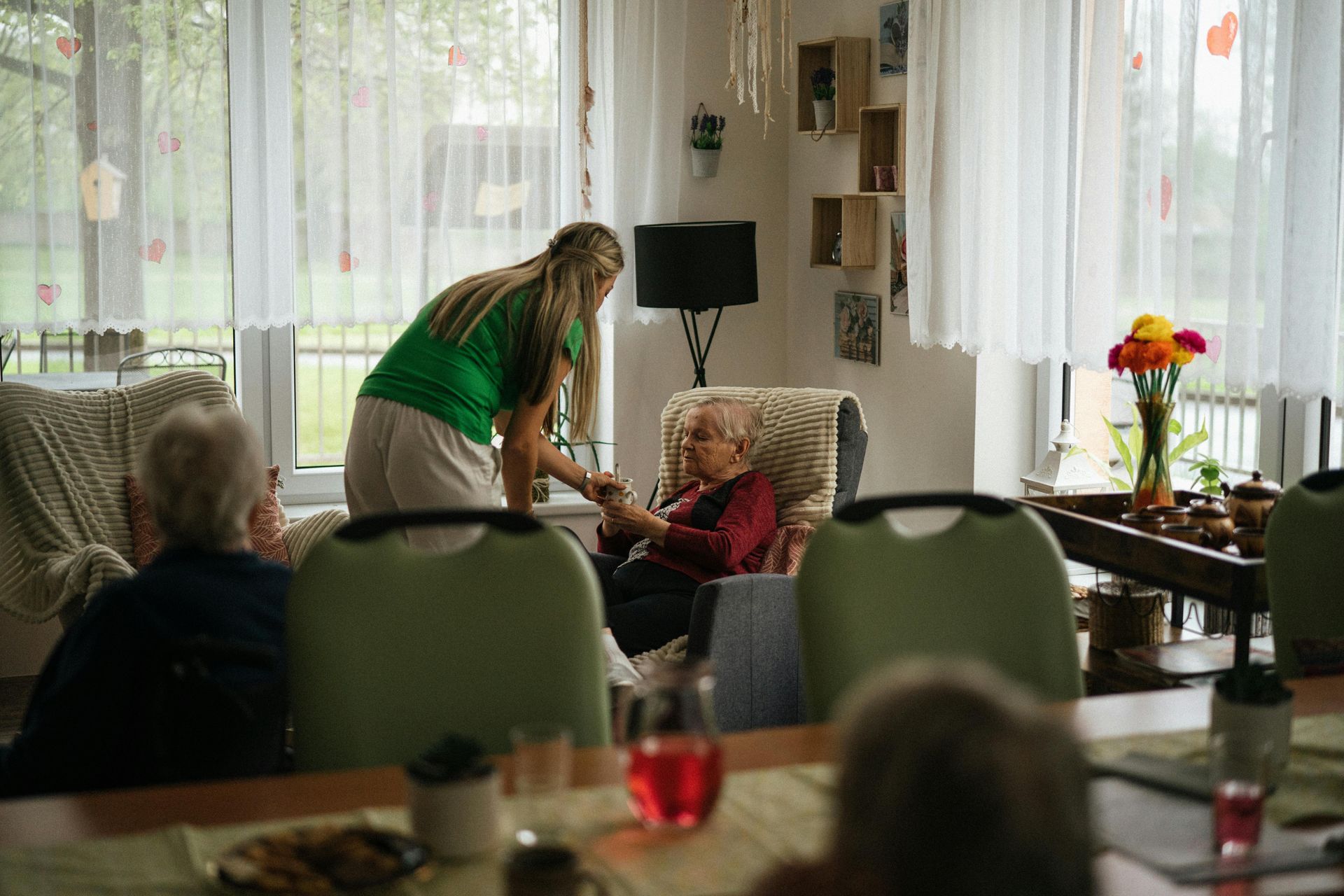 Eine Frau in einem grünen Oberteil hilft einer älteren Person im Wohnzimmer.