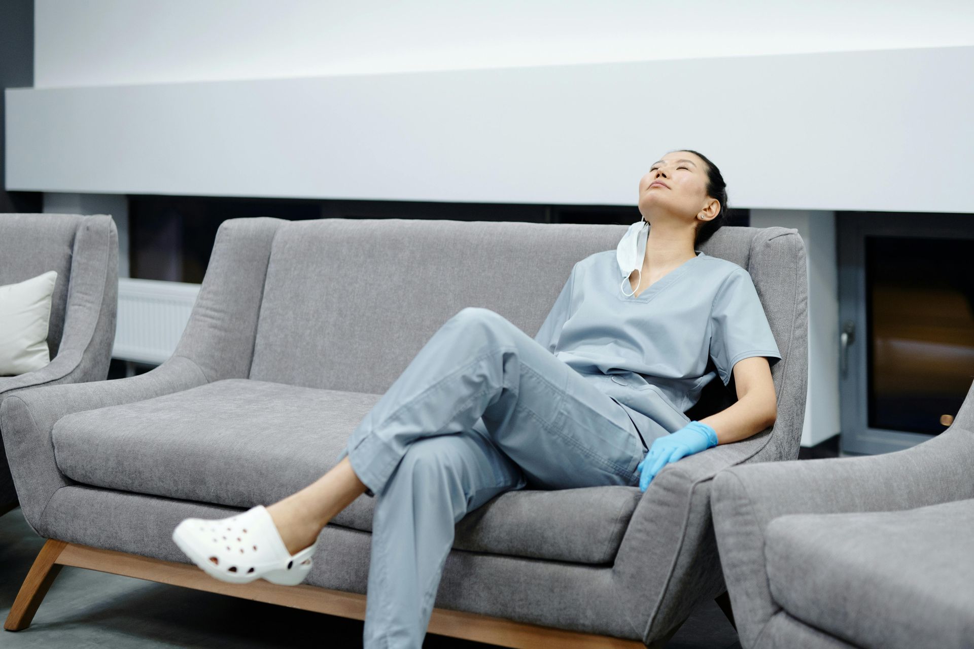 Eine müde Person in OP-Kleidung ruht sich in einem Innenraum auf einem grauen Sofa aus, die Maske um den Hals.