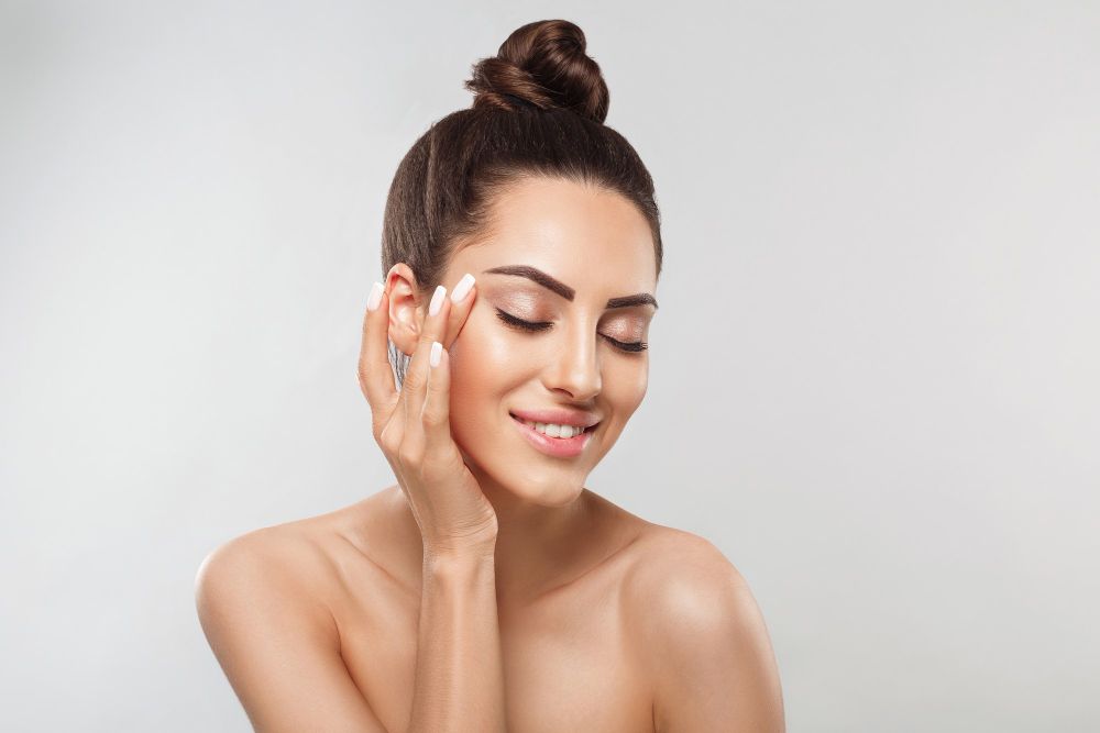 Smiling woman with hair in a bun gently touching her face against a light gray background