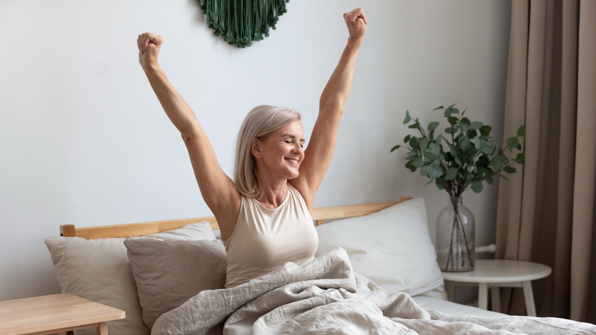 Person sitting up in bed, stretching with arms raised in a bright, cozy bedroom