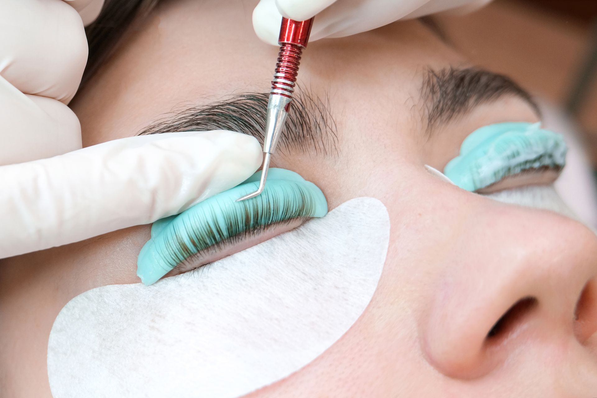 Eyelash extension being applied with tweezers to a client’s closed eye, with under-eye pads in place