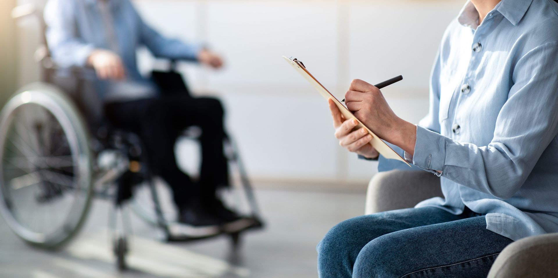 Person in blue shirt taking notes while speaking with a wheelchair user in a bright room