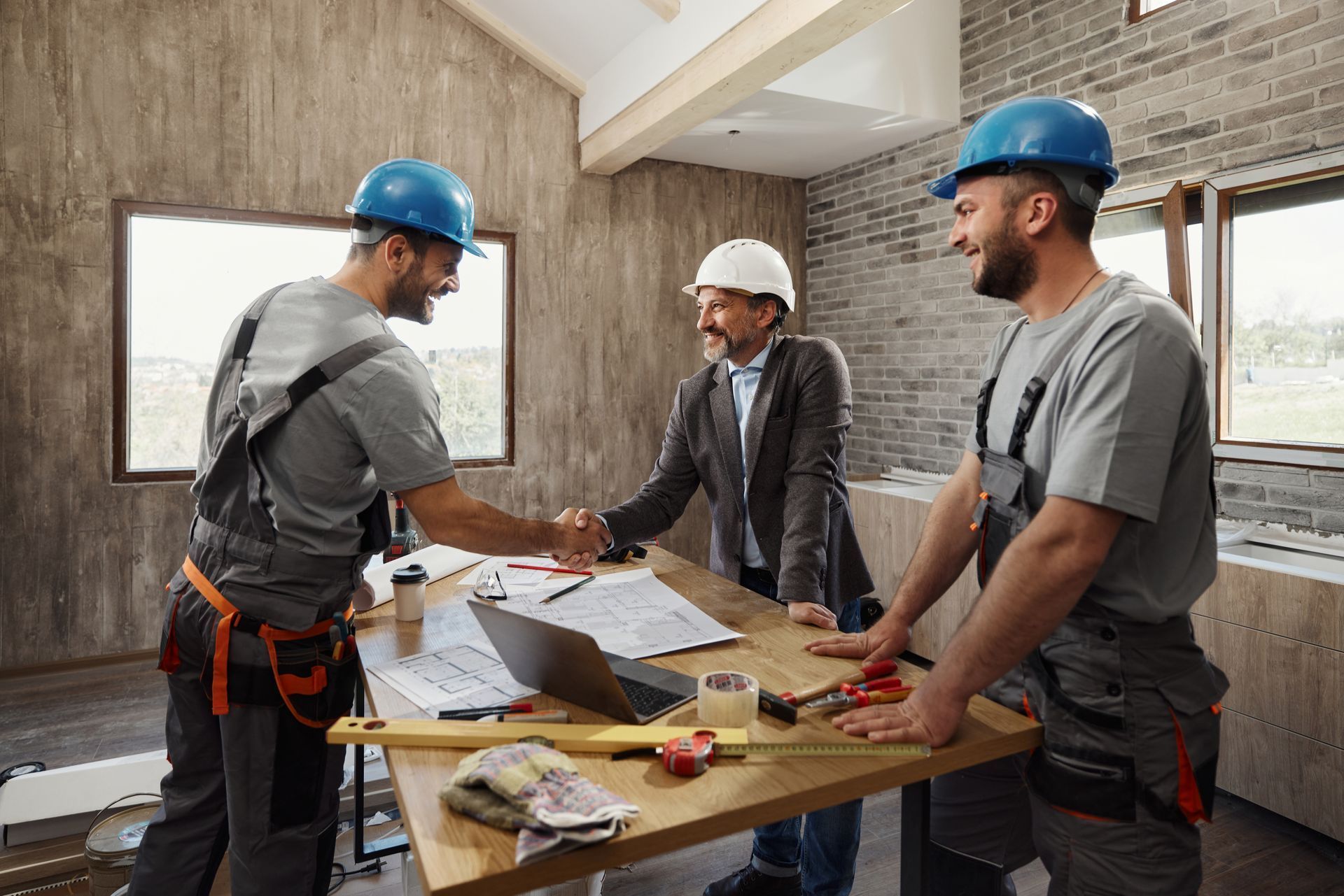 Three construction workers in hard hats review plans around a table in a renovation site.