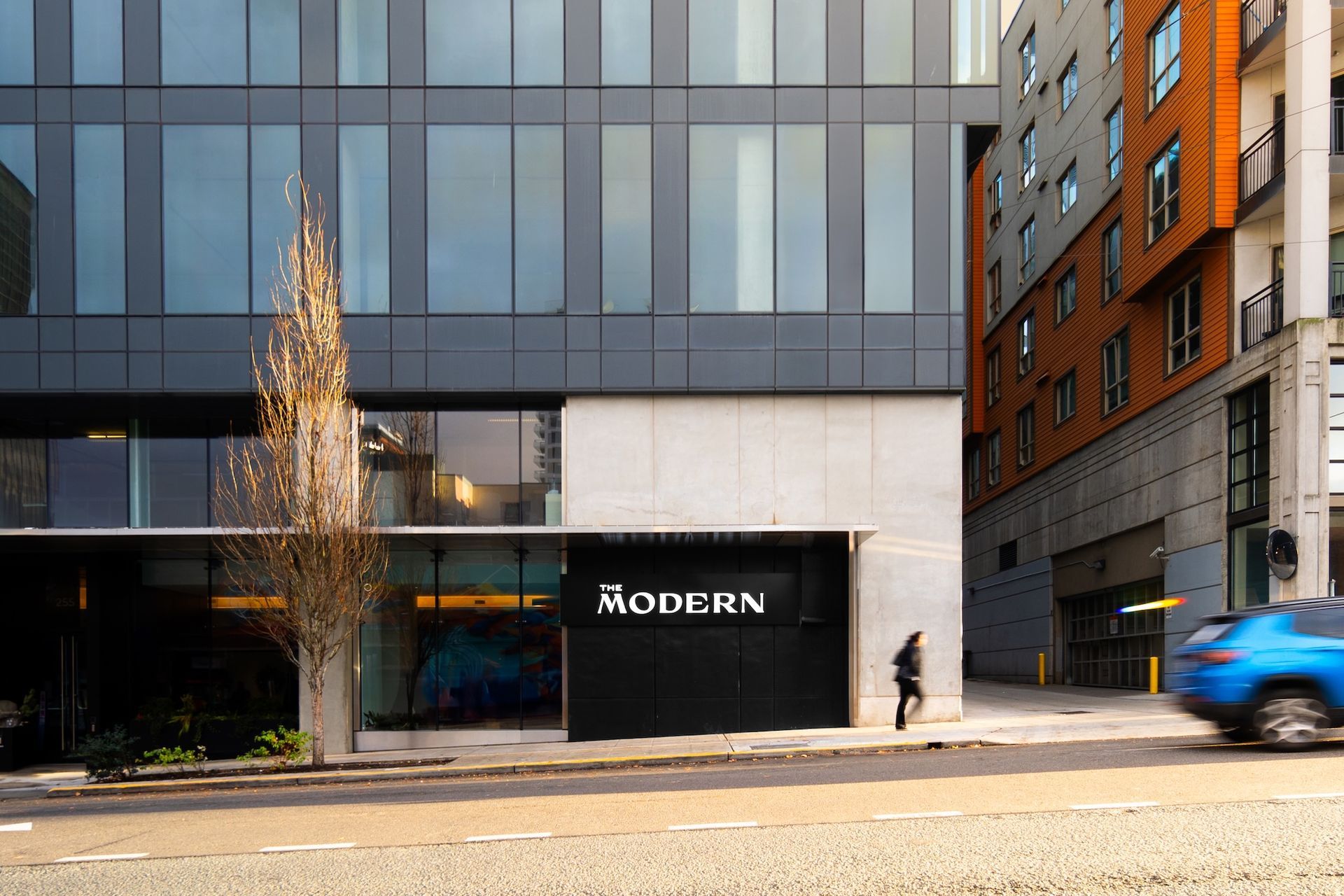 Modern building facade with MODBRN sign, a lone pedestrian, and a blue car passing on the street