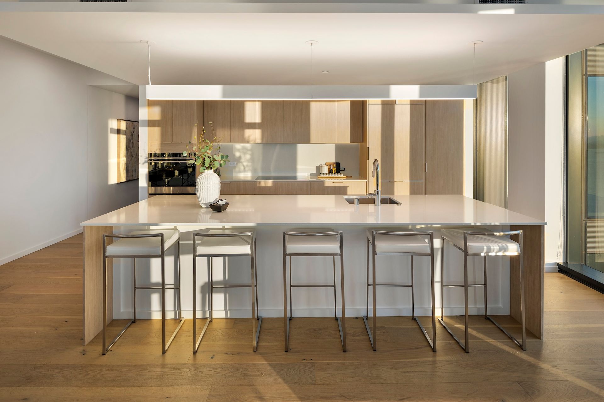 Modern kitchen with a large marble island, bar stools, wood floors, and floor-to-ceiling windows.