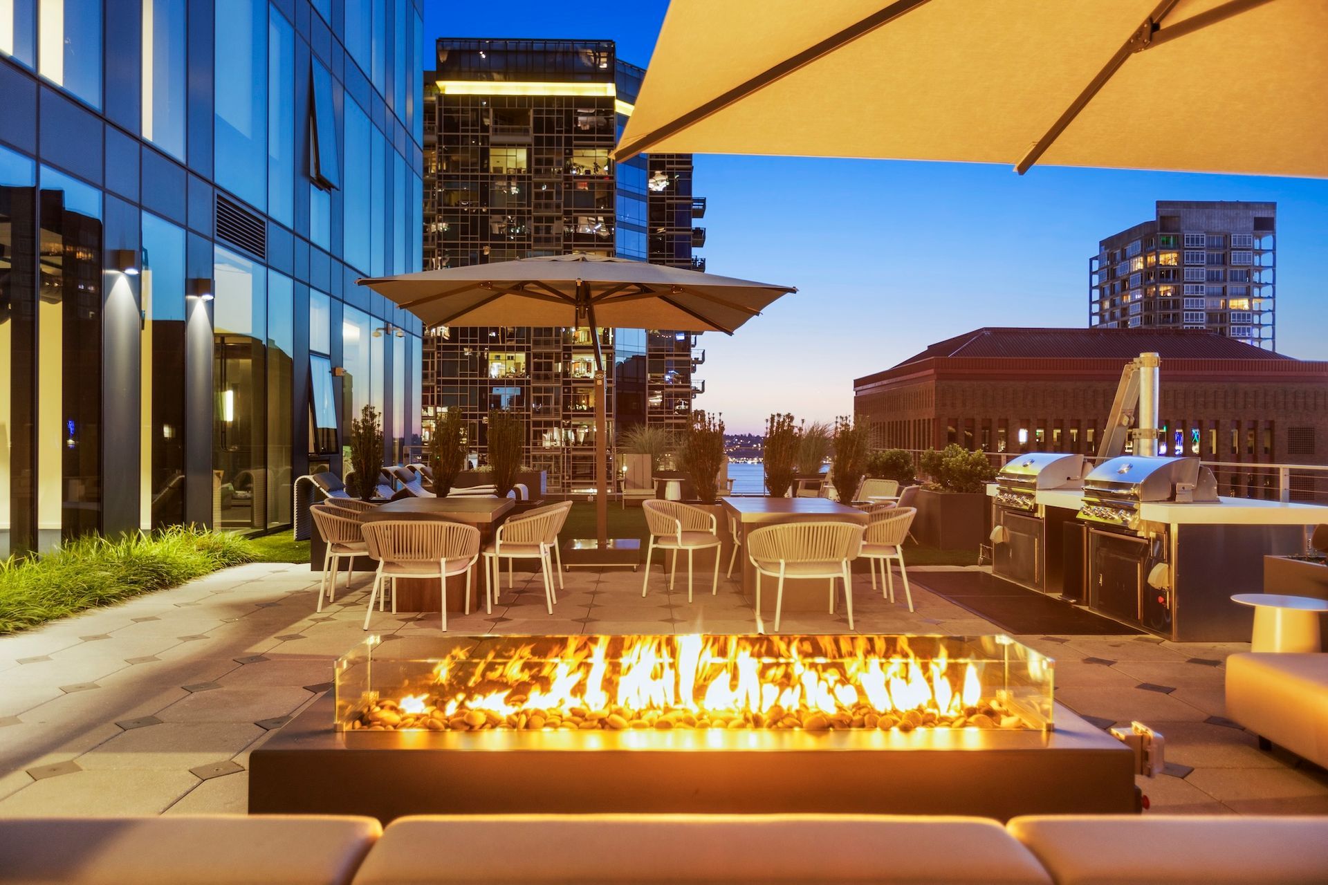 Rooftop patio at dusk with fire pit, outdoor dining tables, umbrellas, and city skyline in the background.