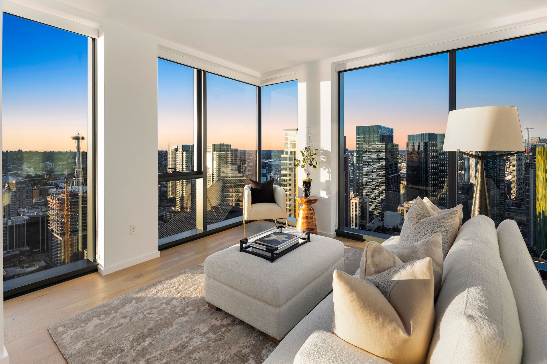 Modern living room with floor-to-ceiling windows, city skyline views, and neutral-toned sofa and ottoman.
